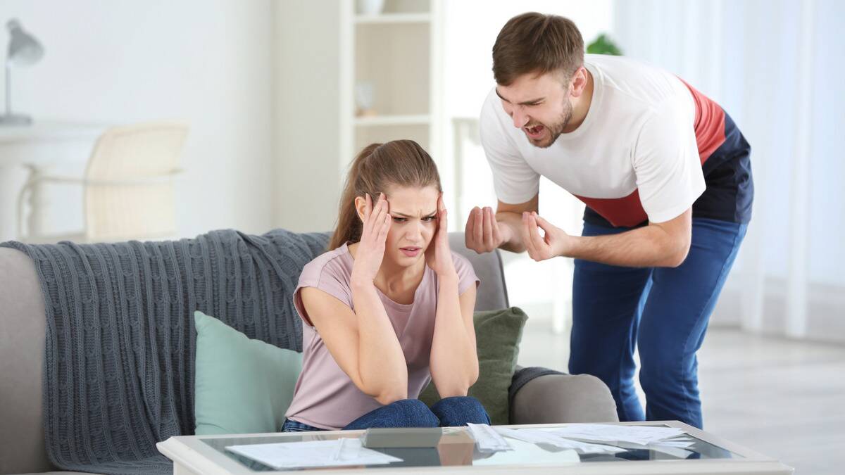 A woman sitting on her couch with her head in her hands, looking confused, while her boyfriend stands next to her, bent down to shout at her.