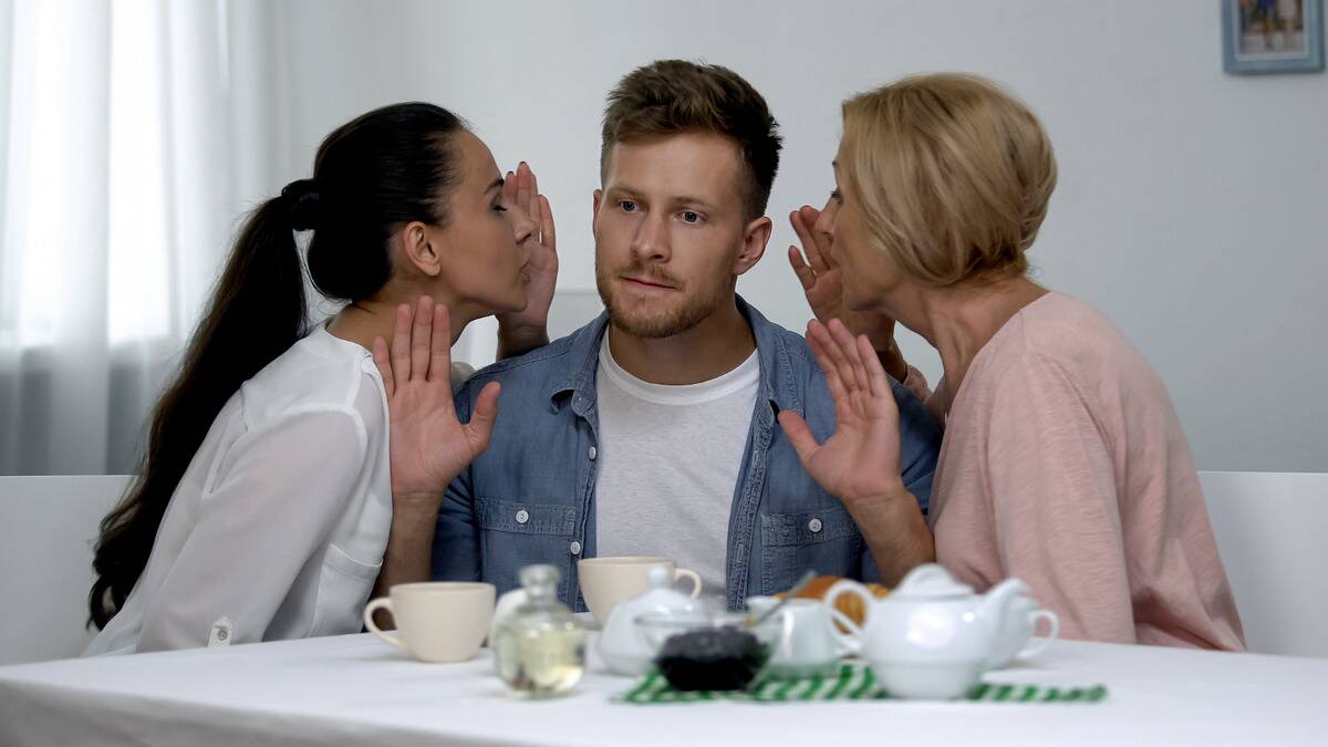 A man sitting at a table, his girlfriend on one side, his mom on the other, both whispering into his ear while he tries to get them to stop.