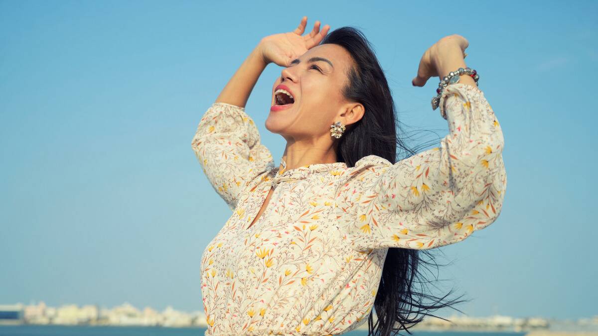 A woman standing outside, arms tossed in the air, mouth open as she shouts joyfully.