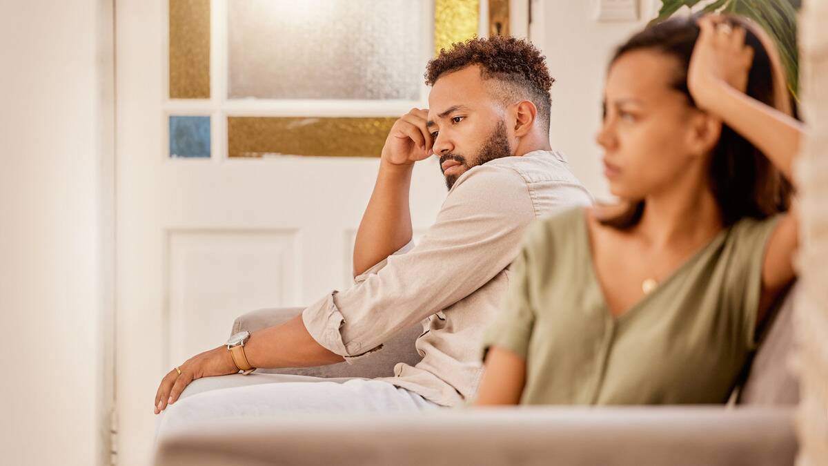 A man and woman sitting next to each other on a couch, distant, not looking at each other, both looking sad, dejected, and annoyed.