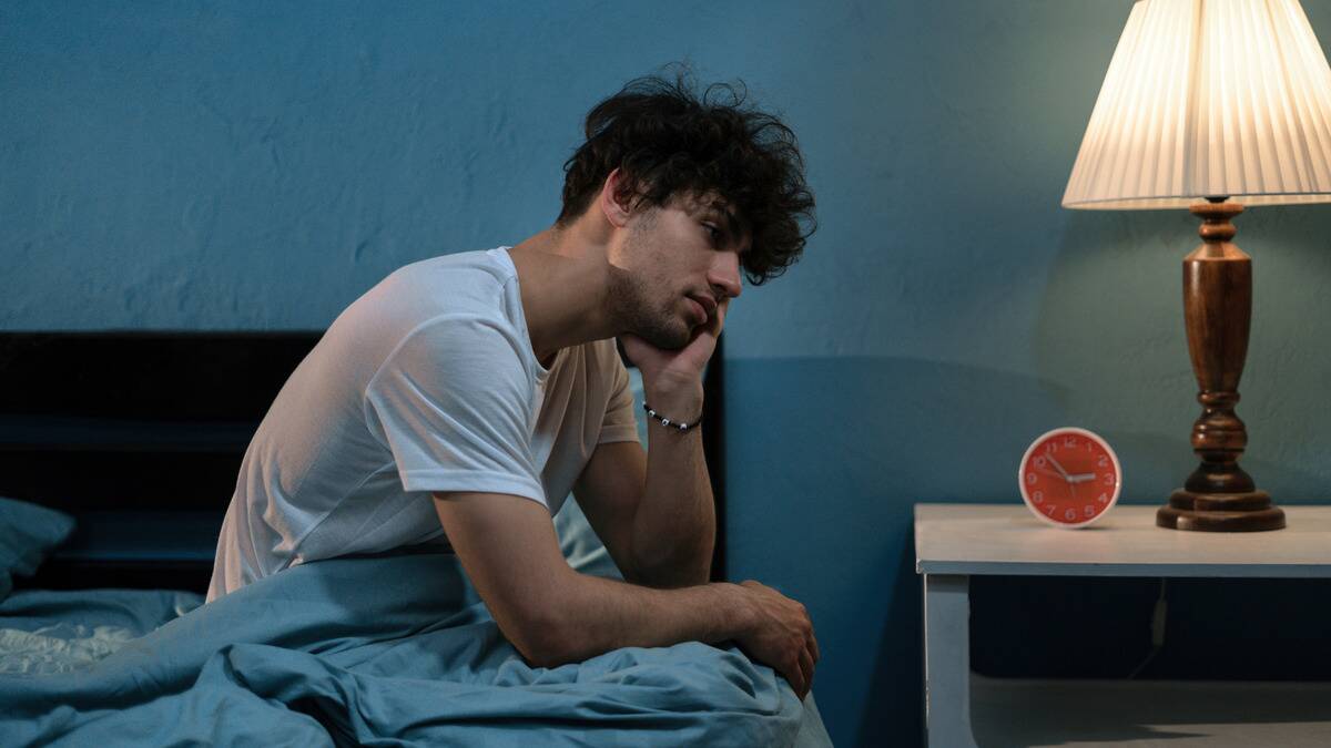 A man sitting up on the side of his bed, one cheek resting on his hand, looking sad.