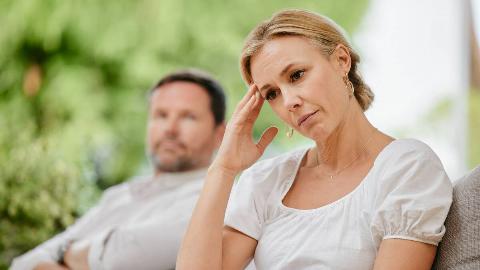 A woman looking stressed, hand to her temple, while a man in the background looks at her disapprovingly with his arms crossed.