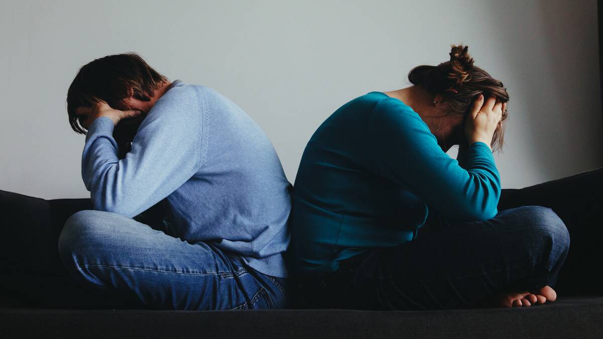 A couple sitting back to back, legs crossed, both leaning their head forward into their hands.