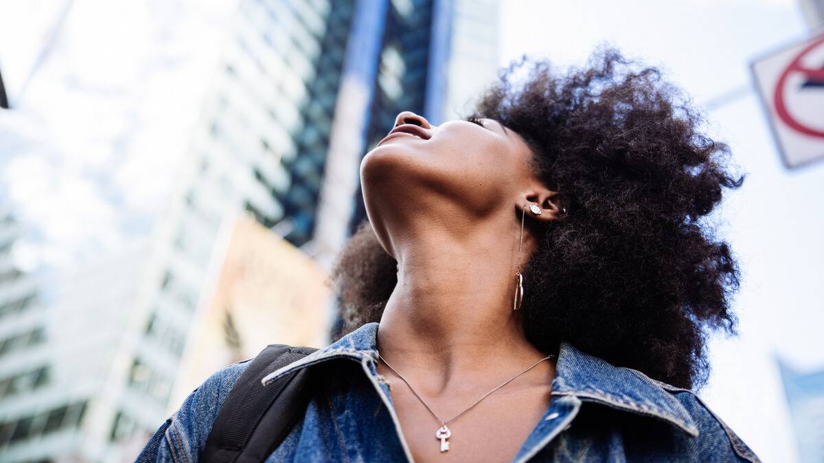 A low angle shot of a woman from the shoulders-up standing on a city street, looking up at the sky.