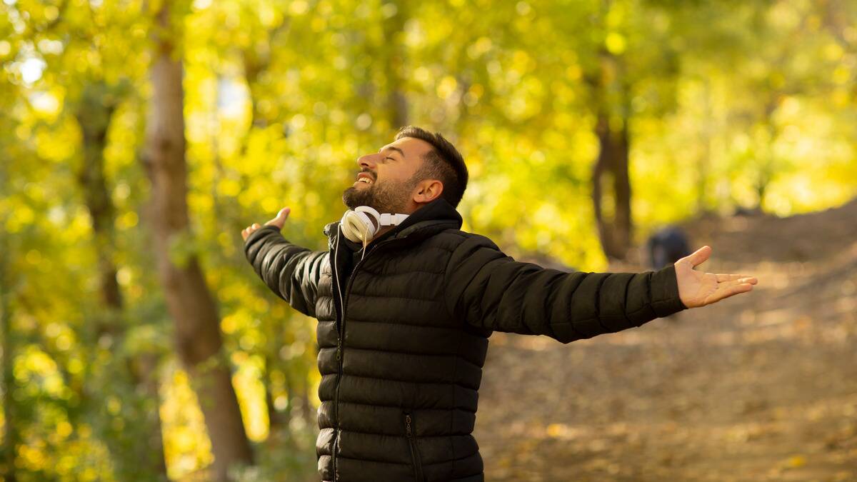 A man standing outside among the trees, arms outstretched, eyes closed, smiling.