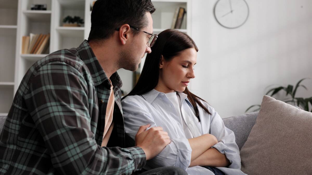 A couple sitting side by side on a couch, the woman with her arms crossed and looking away, the man with a hand on her arm, clearly apologizing.