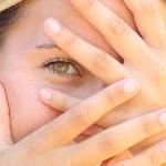 A woman hiding her face behind her hands, one eye peeking out from between her fingers.