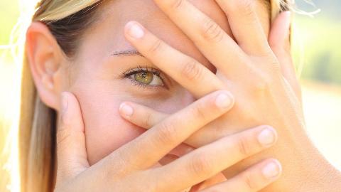 A woman hiding her face behind her hands, one eye peeking out from between her fingers.