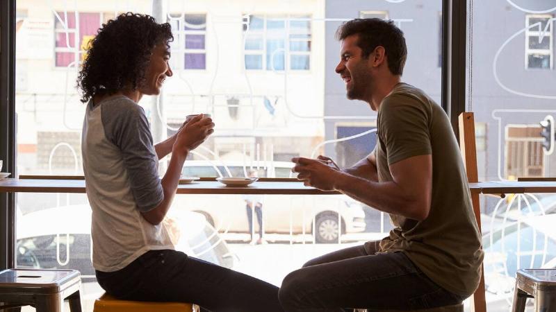 A man and a woman sitting side by side at a cafe counter-style table, both turned toward each other, smiling and laughing as they chat.