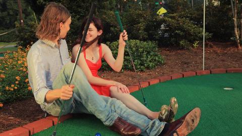 A man and a woman sitting side by side on the side wall of a mini-putt course.