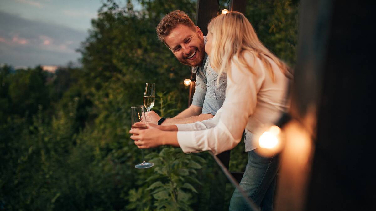 A man and a woman standing side by side as they lean on a railing, talking to one another, each holding a glass of champagne. 