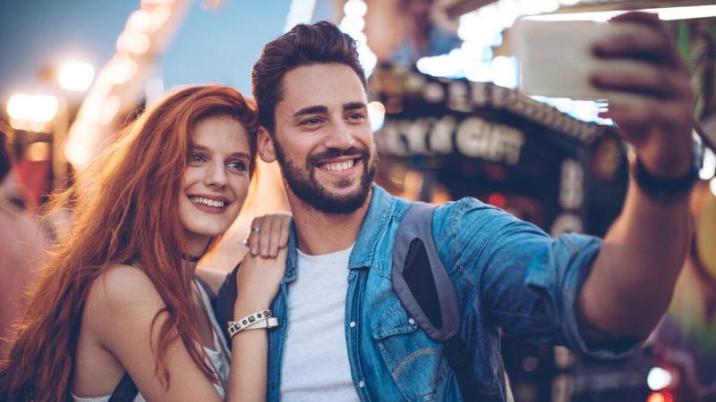 A man and a woman both posing for a selfie at a fair, the man holding his phone out while the woman leans against his shoulder, both smiling.