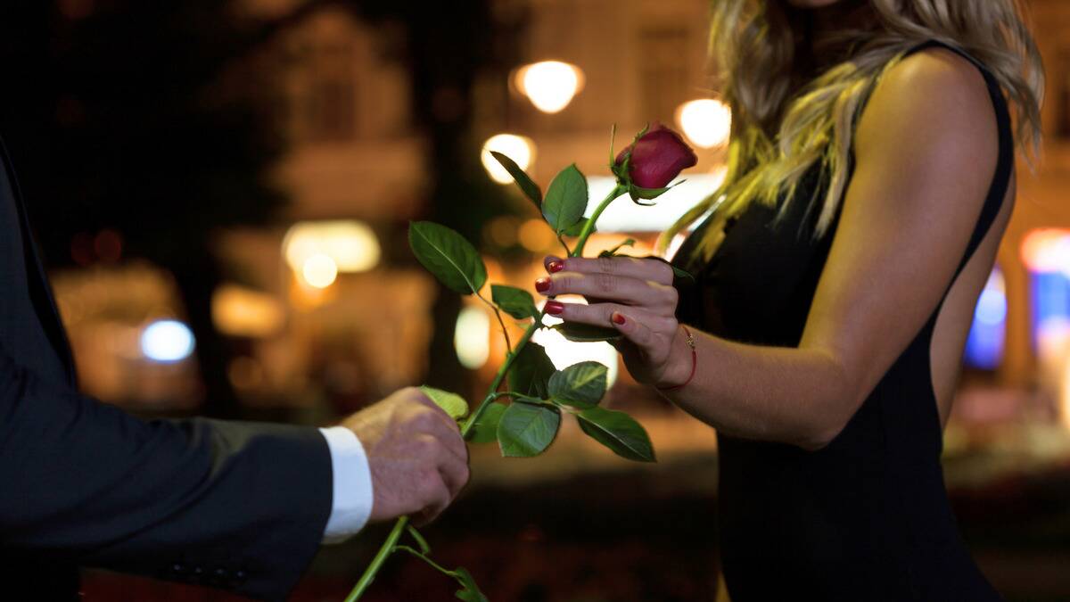 A close shot of a man handing a woman a rose on a night-time street, both dressed fancily.