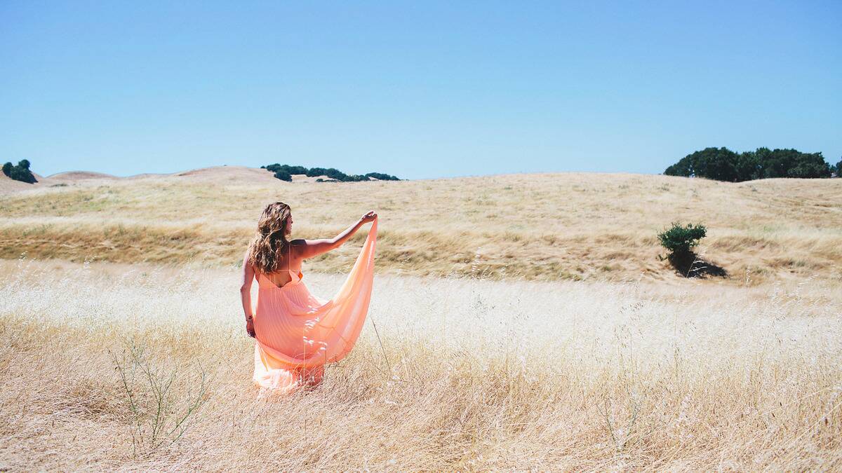 A woman standing in a field of tall grass, holding up one half of her long, flowey dress into the wind.