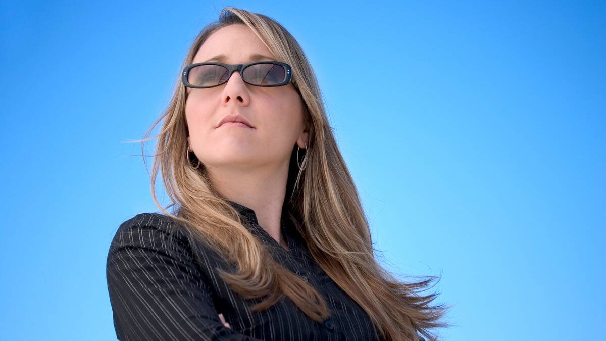 A woman with her arms crossed and sunglasses on as she stands beneath a clear bright blue sky.