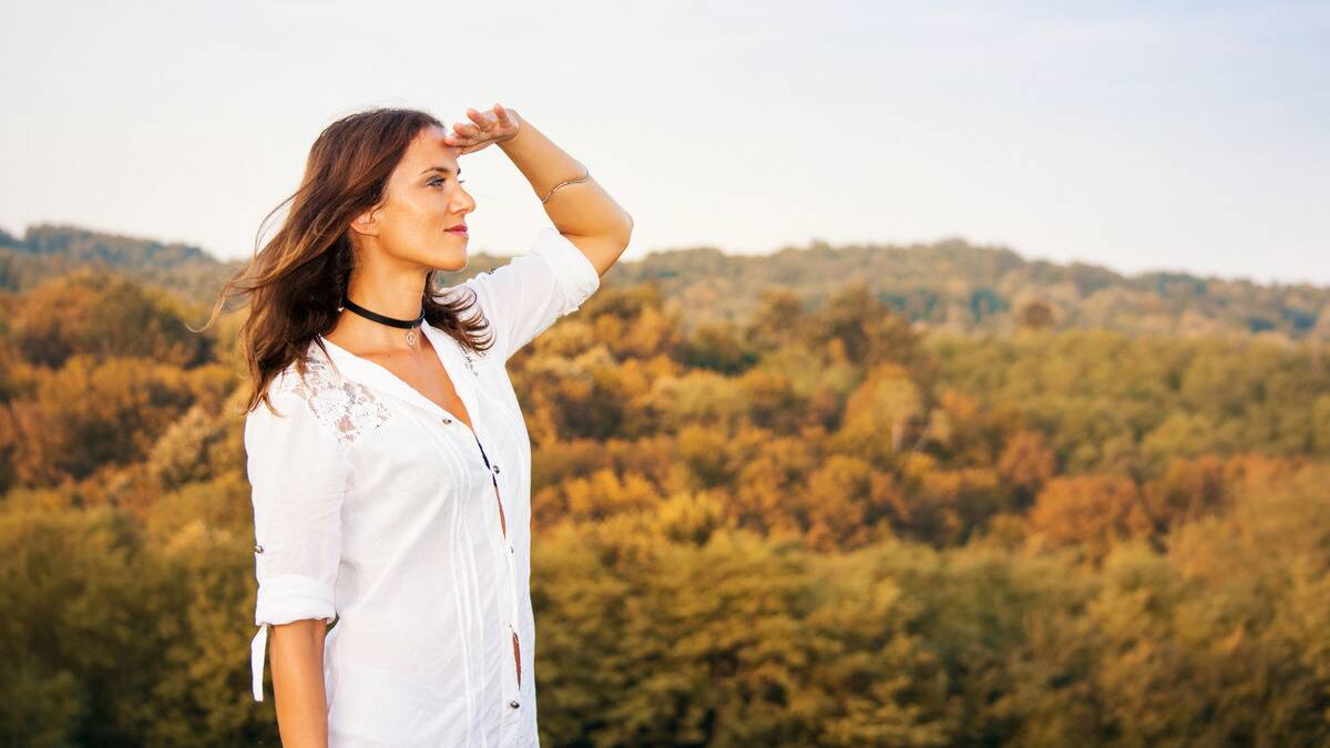 A woman standing outdoors in front of a hill range, a hand up to her brow, looking forward.