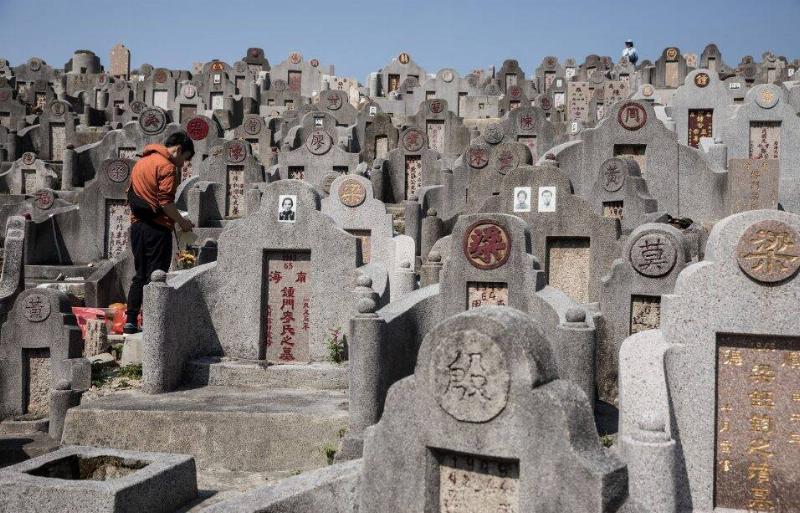 A crowded Chinese cemetery, uneven rows full of headstones with altars for offerings. On the left, one person stands in front of one of those headstones with their head bowed.