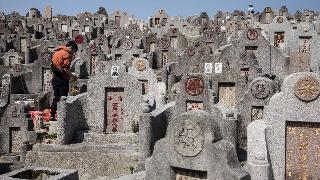 A crowded Chinese cemetery, uneven rows full of headstones with altars for offerings. On the left, one person stands in front of one of those headstones with their head bowed.