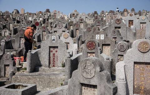 A crowded Chinese cemetery, uneven rows full of headstones with altars for offerings. On the left, one person stands in front of one of those headstones with their head bowed.