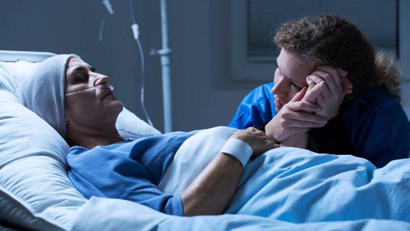 A woman laying still in a hospital bed, clearly in critical condition, as a woman sits next to the bed, holding the patient's hand in both of her own, leaning her face against it.