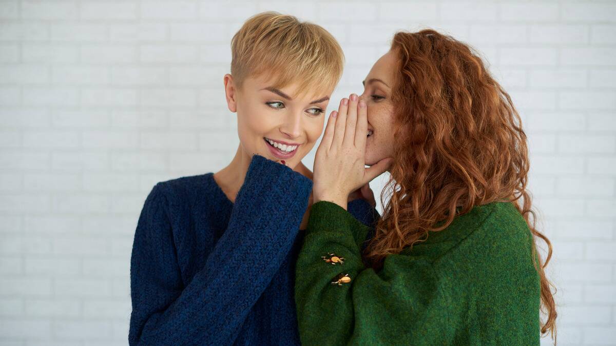 Two women smiling as one whispers a secret into the other's ear.