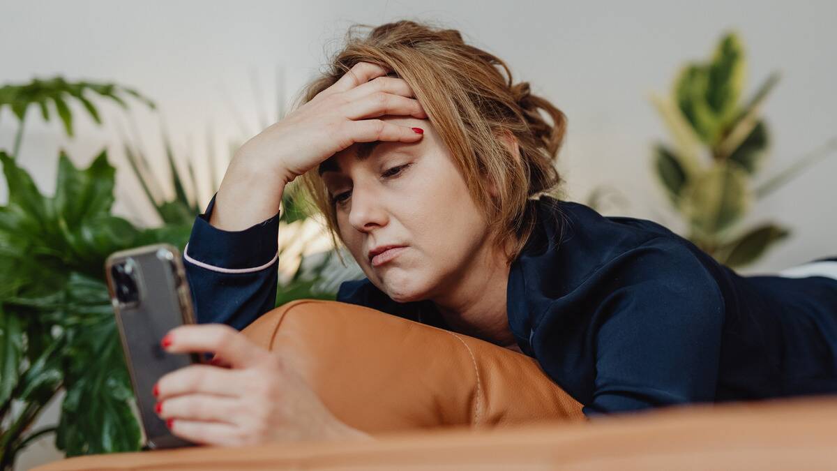 A woman laying on her stomach with a hand on her forehead as she looks at her phone, sporting a sad expression.