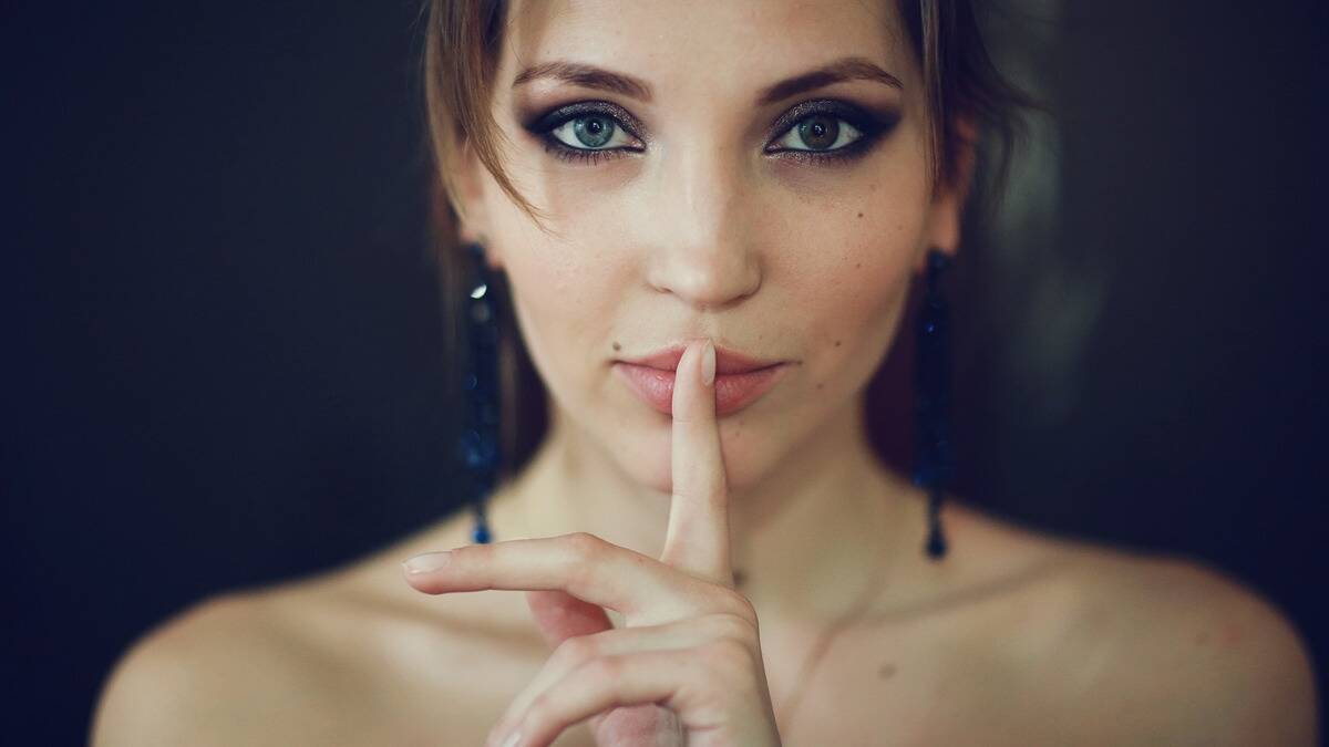 A woman in glam makeup with a finger over her mouth.