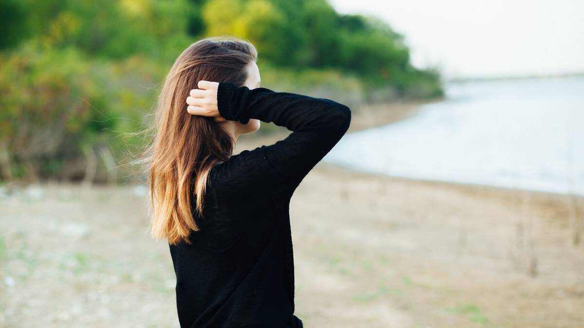 A woman standing on the beach, looking out toward the water, one arm raised to push her hair behind her ear.