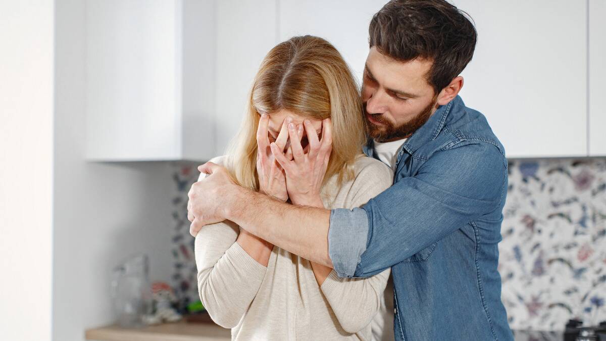 A man hugging a woman from the side as she covers her face with both hands, clearly upset about something.