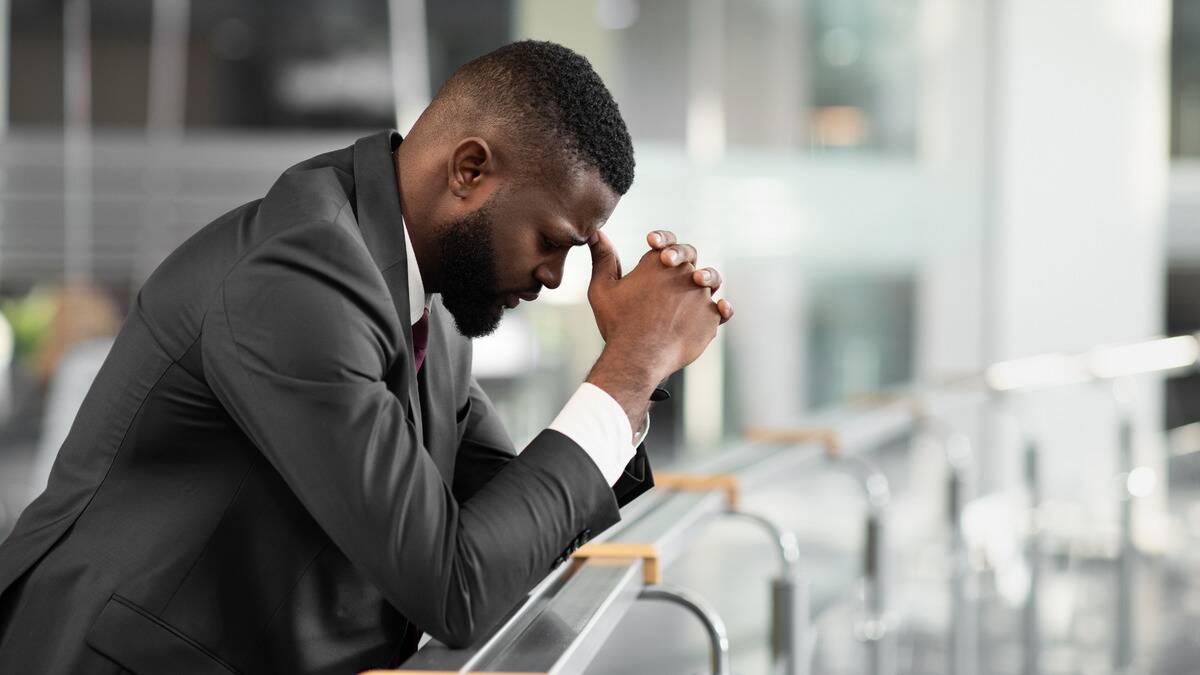 A man in a business suit leaning against a railing in a building, forehead against his folded hands, looking frustrated.