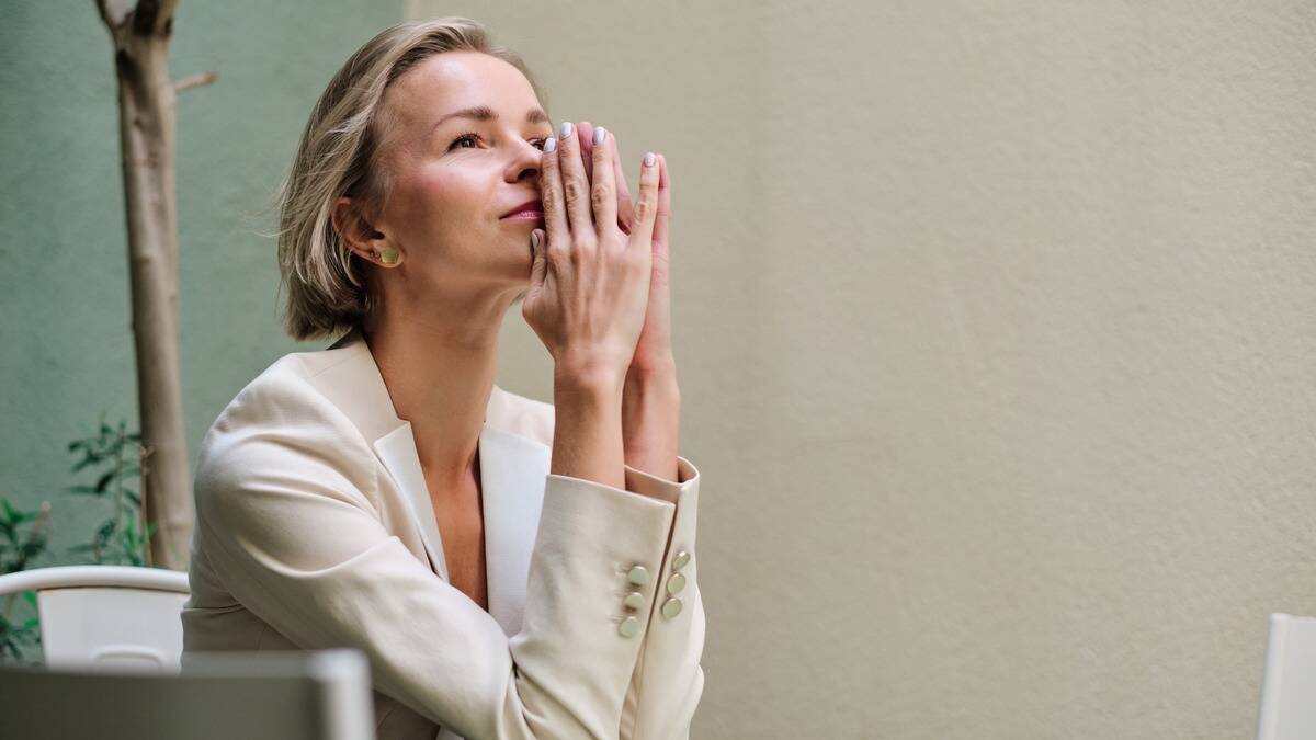 A woman sitting at a table, hands pressed together, pressed against her nose, mouth, and chin.