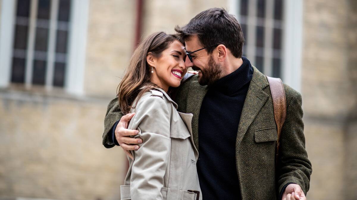 A couple standing close on the street, the man with an arm around the woman, their noses touching, both smiling.