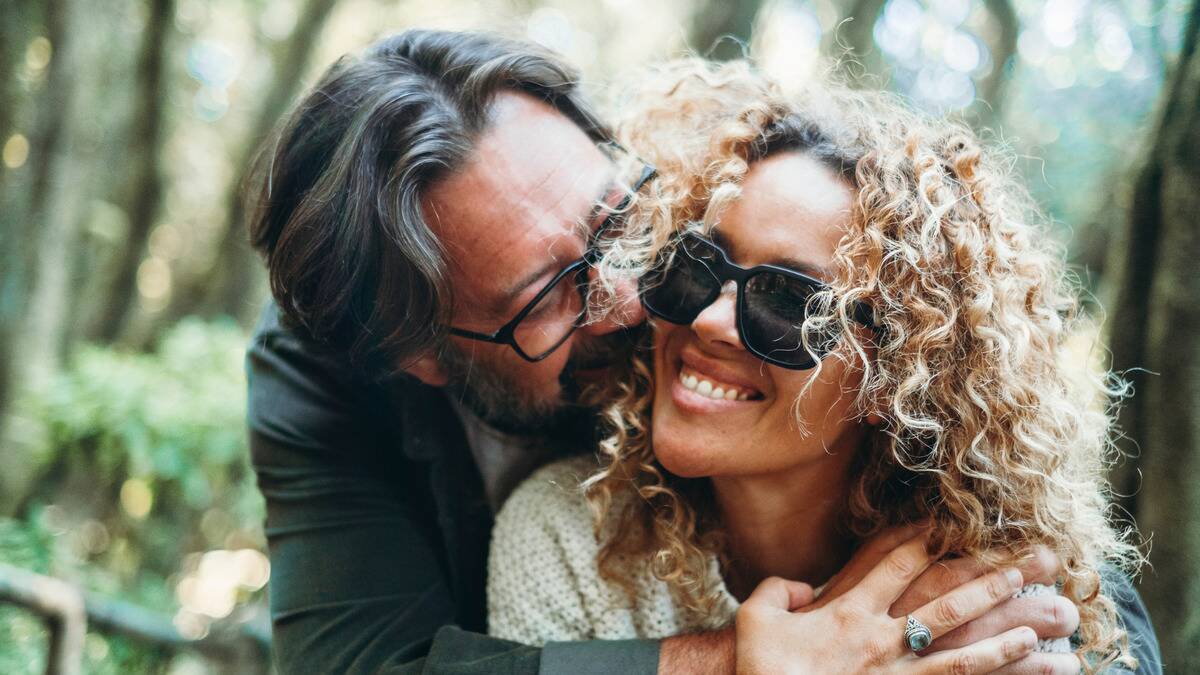 A close shot of a couple hugging close outdoors, the man embracing the woman form behind as they both smile.