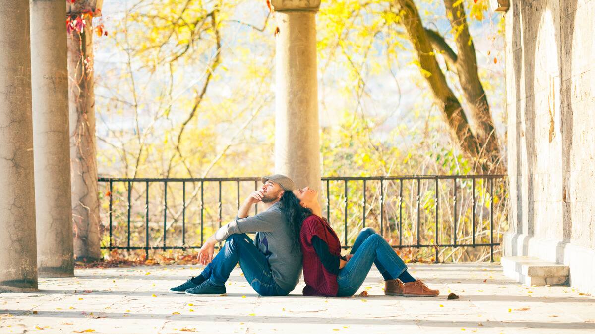 A couple sitting on the stone ground of a terrace, backs to each other, knees bent, both looking tired or stressed.