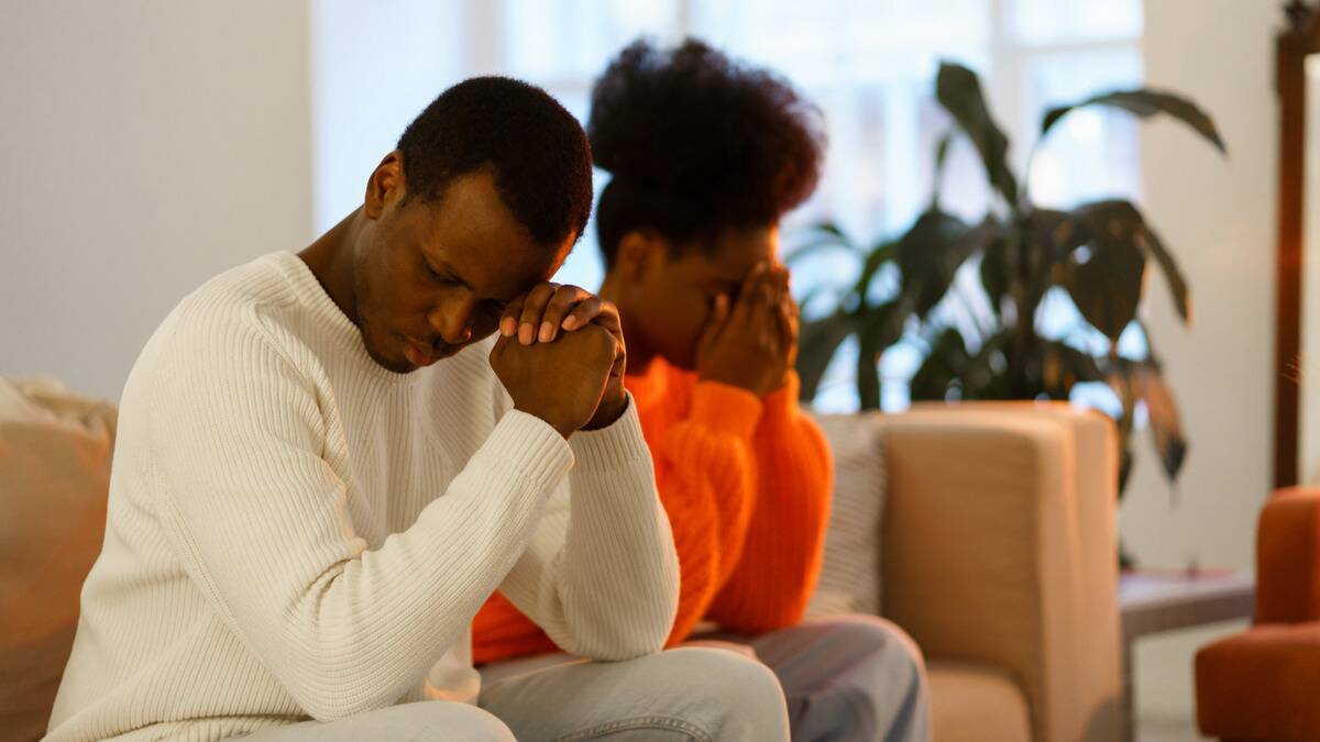 A couple sitting side by side on a couch, both with their elbows on their needs, the man with his hands together leaning his temple against them, the woman with both hands covering her face.