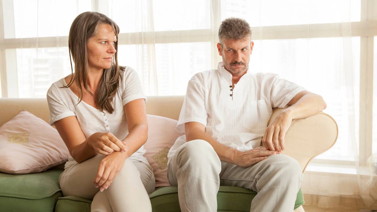 A couple sitting side by side on a couch but leaning away from each other, both looking at each other awkwardly.