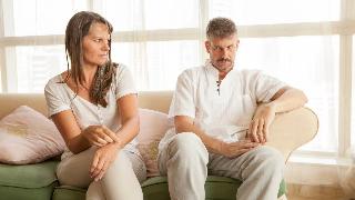 A couple sitting side by side on a couch but leaning away from each other, both looking at each other awkwardly.