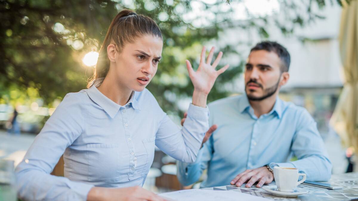 A woman angrily dismissing her boyfriend while they're out at a cafe, holding a hand up to stop him from speaking.