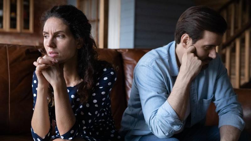 A couple sitting next to each other, but facing away from one another, the woman with her hands folded in front of her chin, the man with his hand on his cheek.