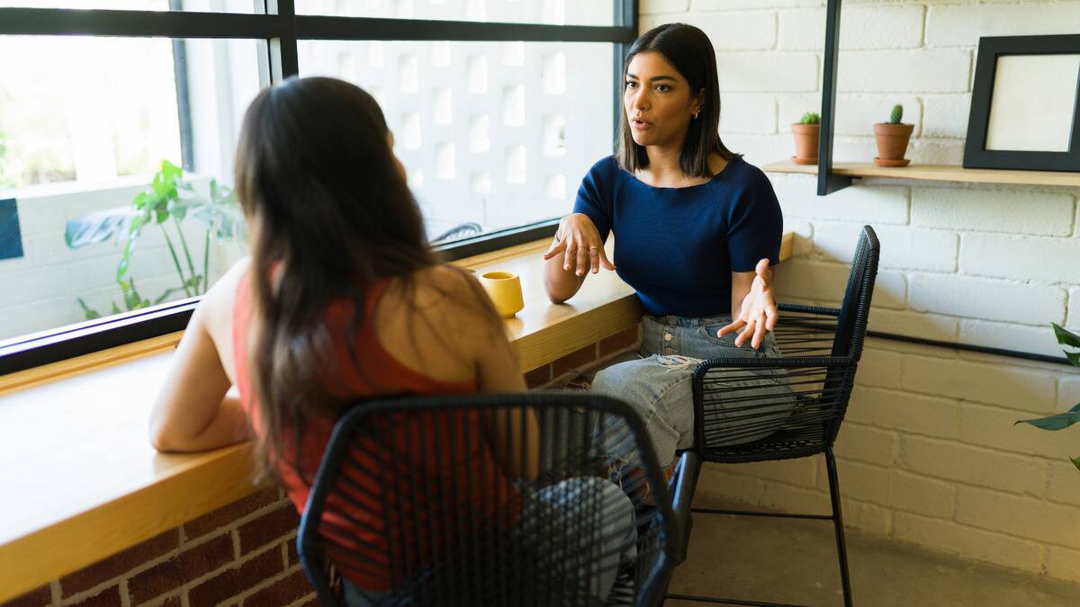 Two friends at a cafe sitting next to each other, turned toward one another, one speaking while the other listens.