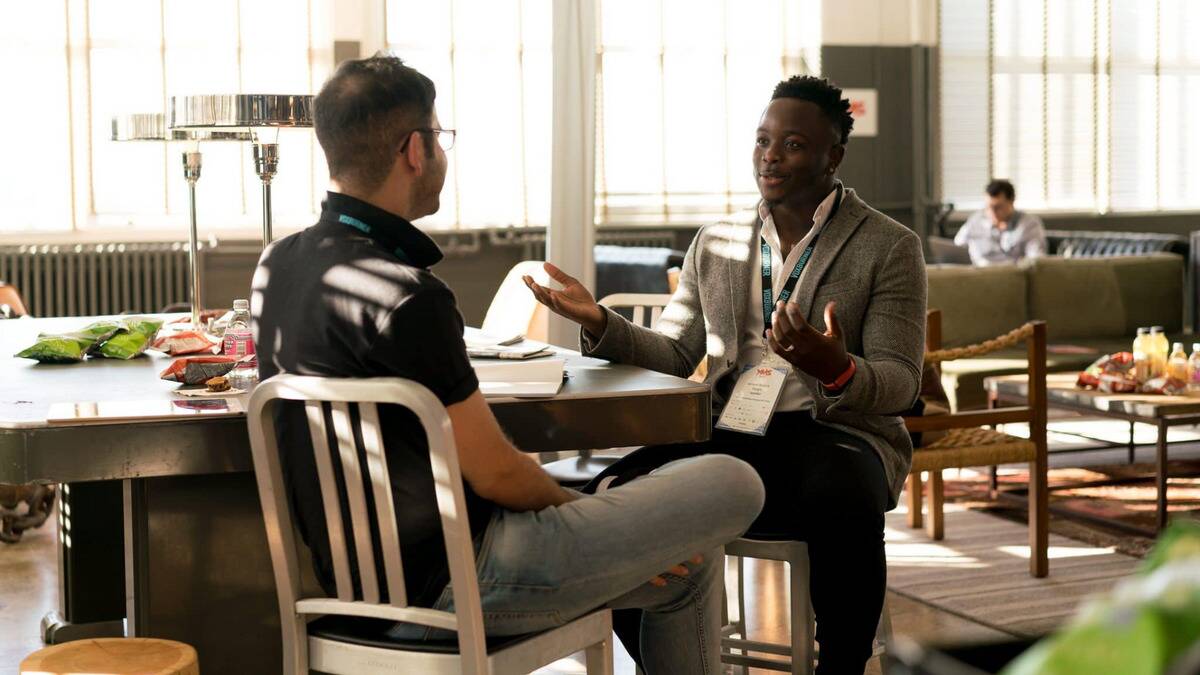 Two men sitting across from one another at an event, one speaking and gesturing with his hands while the other listens.