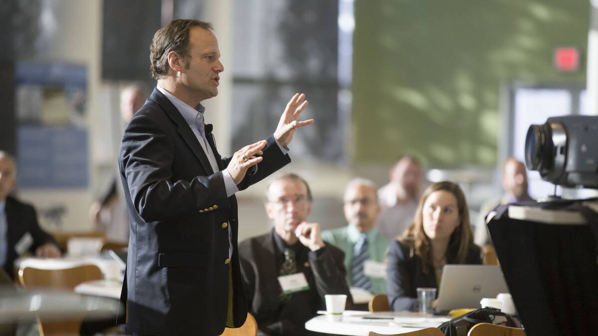 A man confidently giving a presentation to a large group of people, all in business attire.