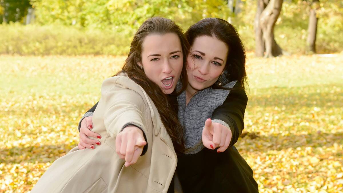 Two women with an arm around one another, pointing at the camera with cocky expressions.