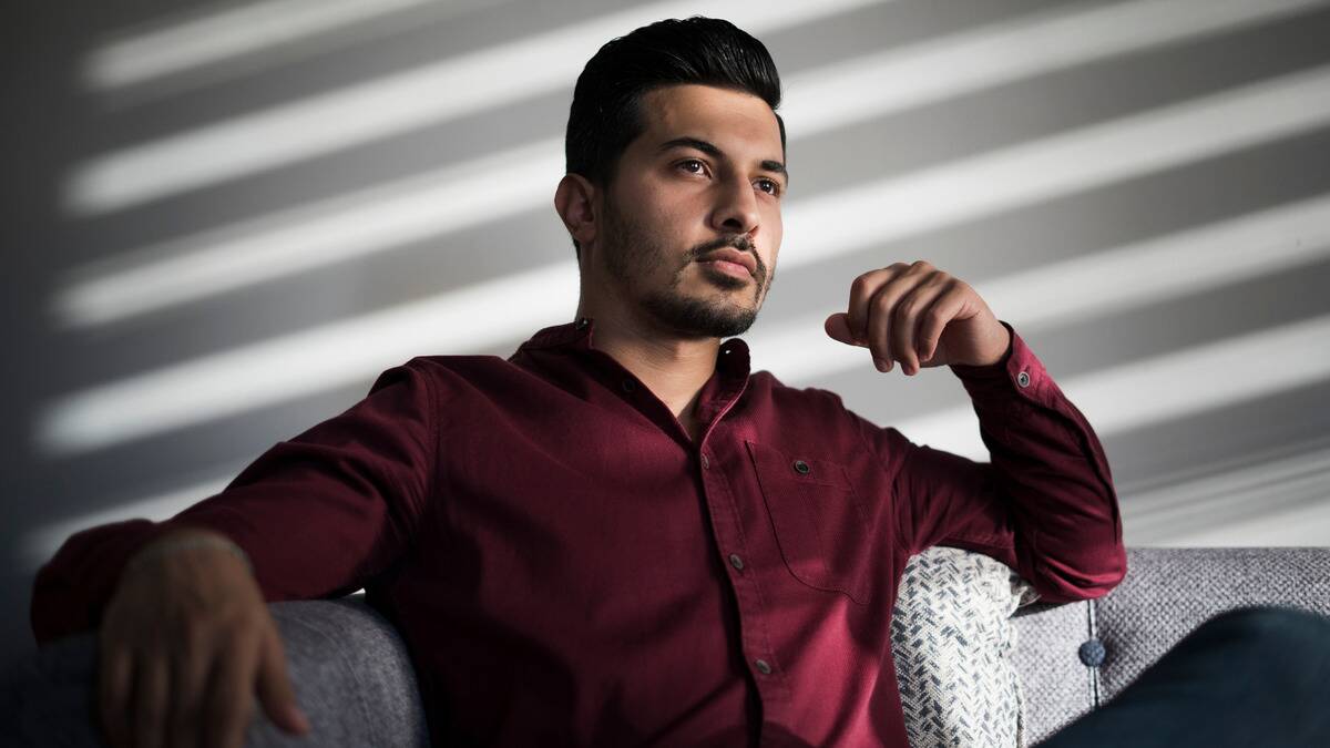 A man sitting on a couch looking out a window that's half covered with blinds, seeming serious.