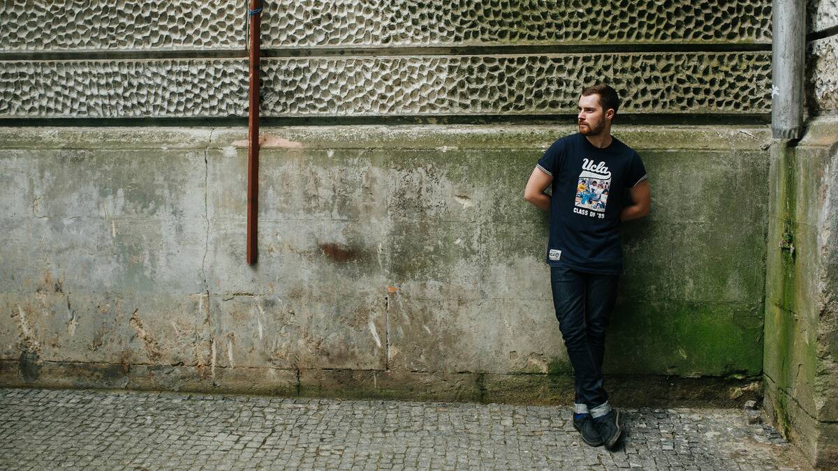 A man leaning against a stone wall in a city, hands behind his back, legs crossed looking off to the side.