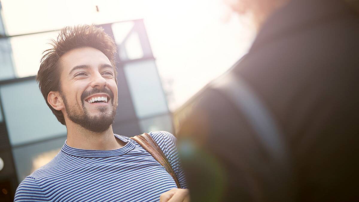 A low angle shot of a man smiling as he meets someone new, both standing outside.