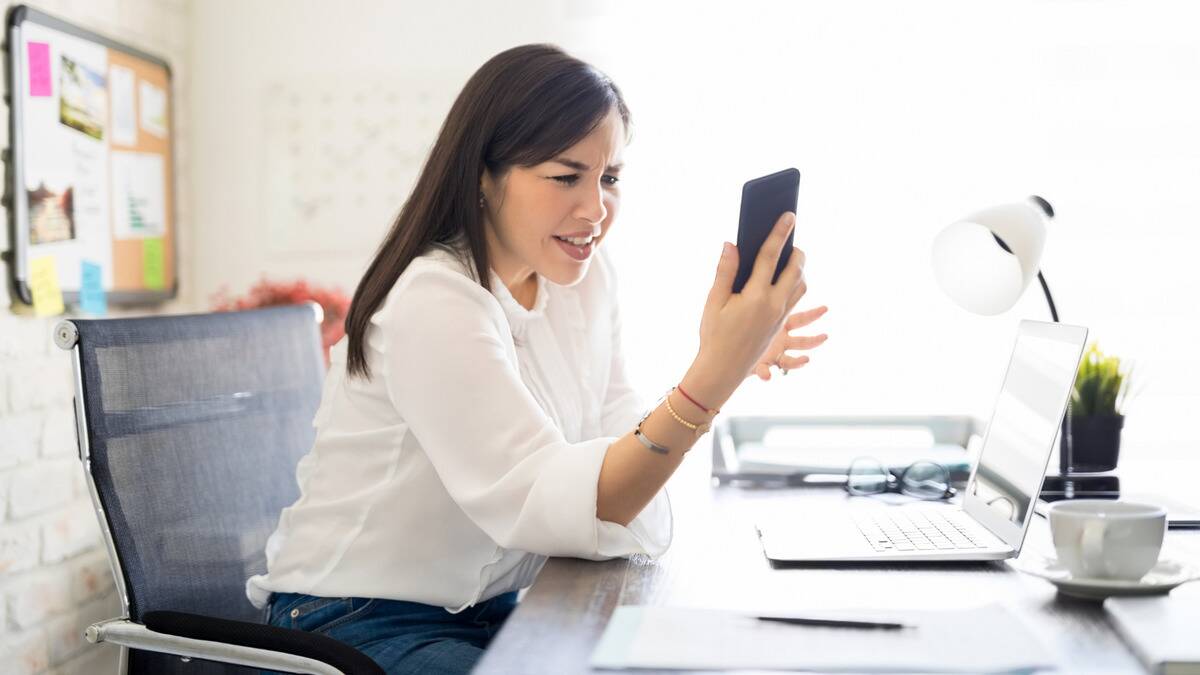 A woman sitting at her office desk looking at her phone, seeming to be complaining.