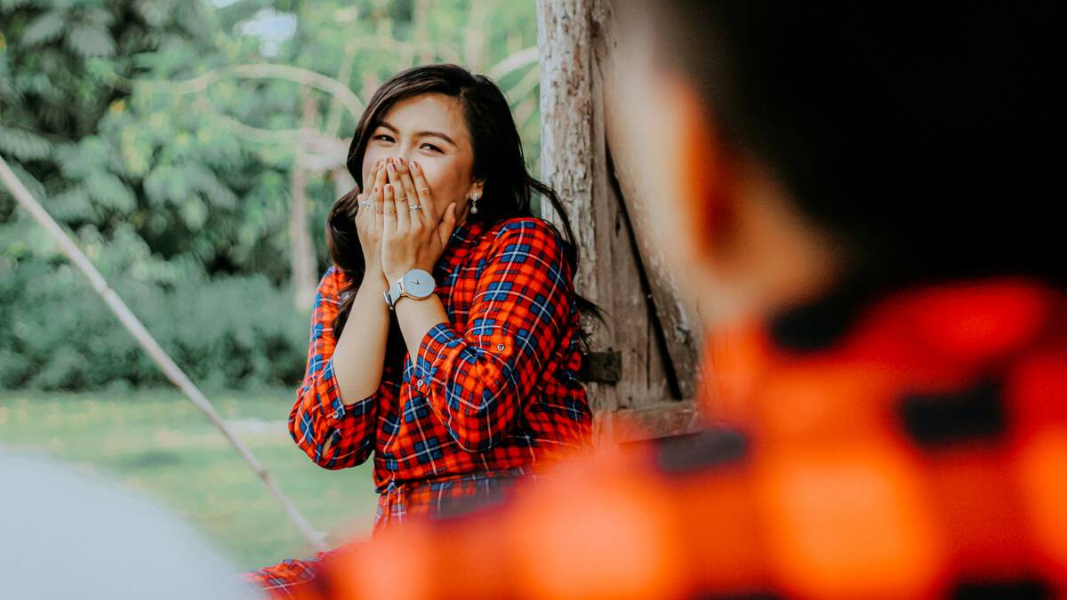 A woman covering her mouth as she laughs at someone whose perspective we're seeing, both standing outside.