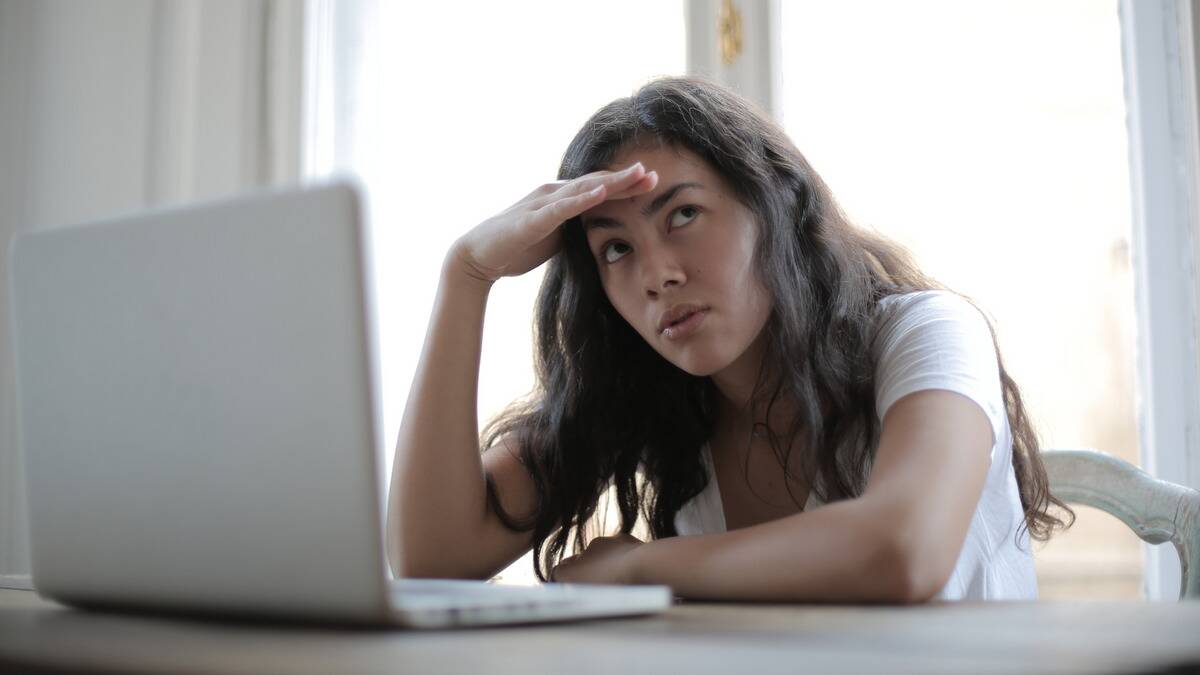 A woman sitting at a table with a laptop in front of her, a hand on her brow bone, looking off to the side with an annoyed expression.