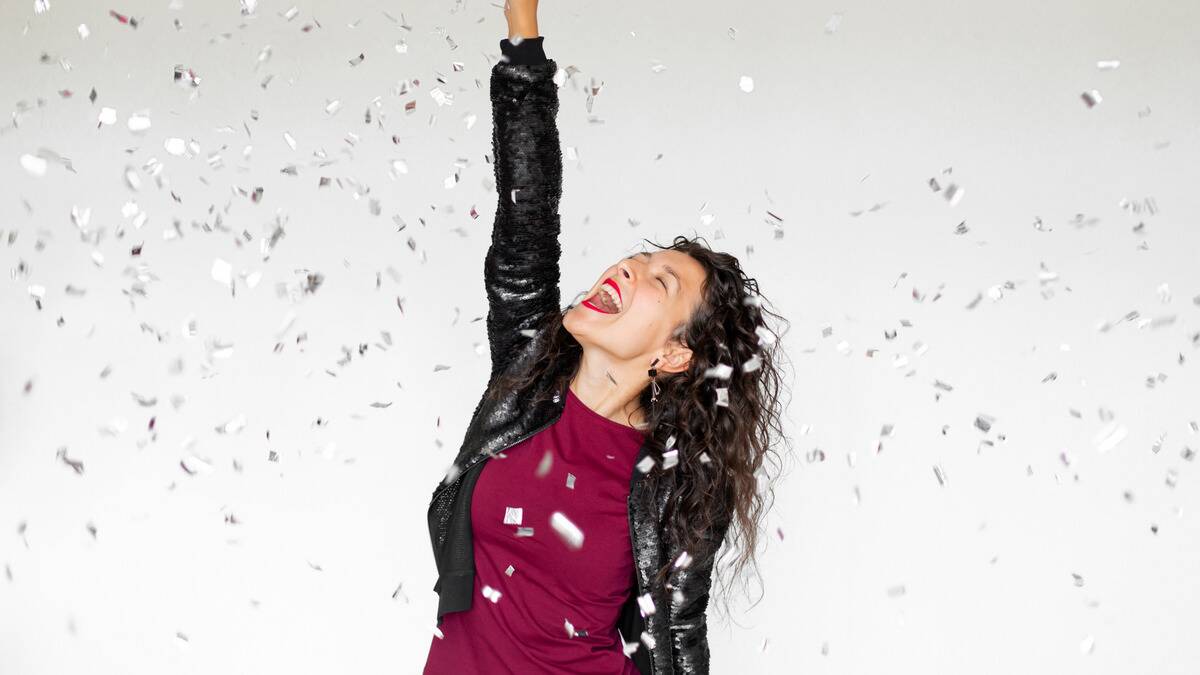 A woman yelling gleefully, eyes closed, looking up, throwing one arm in the air as confetti rains down around her.